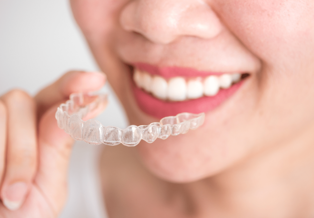 Person holding clear dental aligner, smiling to display whitened teeth against neutral background.