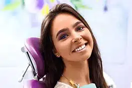 Smiling woman with long dark hair wearing gold necklace in dental office setting.