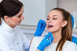 Two dental professionals in clinical setting; one examines patient's teeth wearing blue gloves.