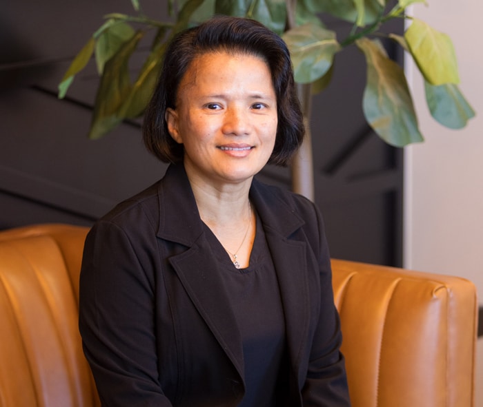 Professional woman in dark blazer seated on orange leather couch with plant behind her.