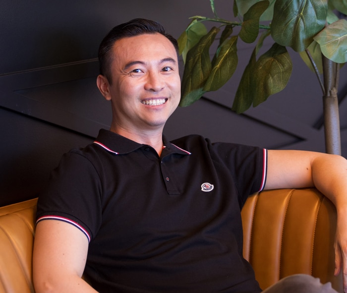 Smiling man in black polo shirt seated on tan leather couch in modern office setting.
