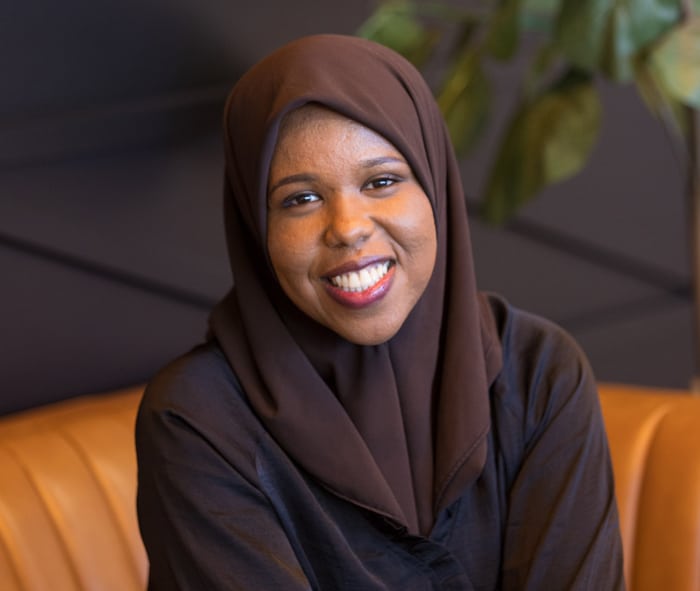 Woman wearing hijab smiling at camera, seated on orange couch in professional dental office setting.