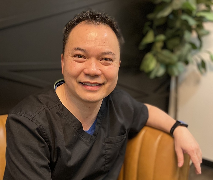 Smiling dental professional in black scrubs seated in modern office with plant backdrop.