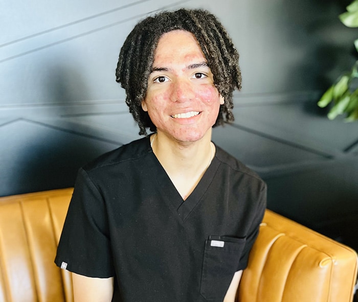 Young man in black scrubs smiling at camera, seated on yellow leather chair in dental office.