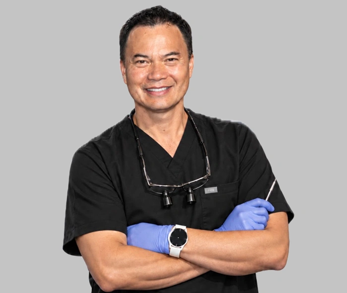 A smiling East Asian male dental professional posing against a solid light gray background with arms crossed, wearing black scrubs and medical loupes around his neck.