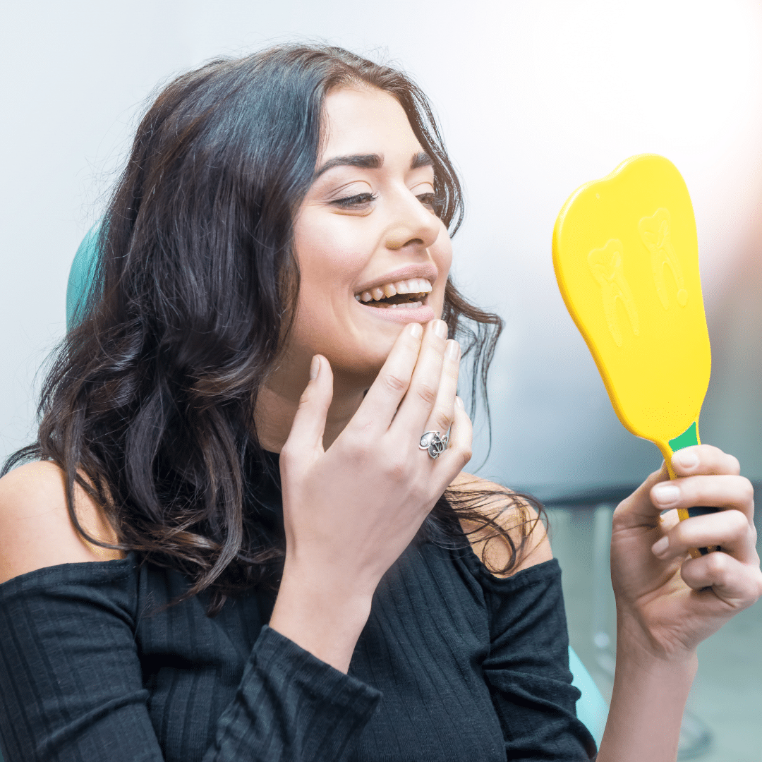Woman holding yellow mirror examining teeth and smiling in dental office setting.